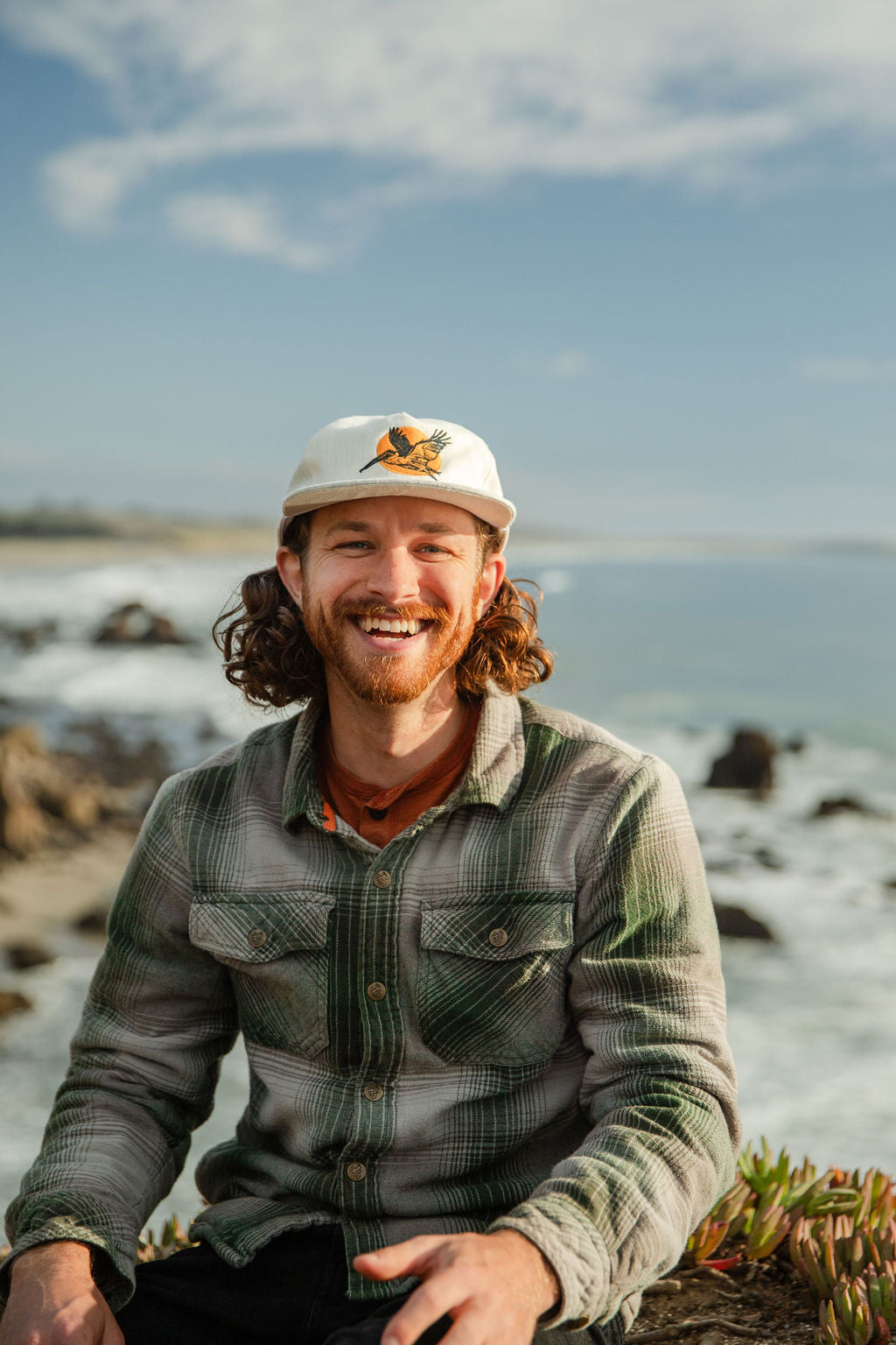 Sam Blake sitting on ocean cliff with hemp pelican hat, smiling